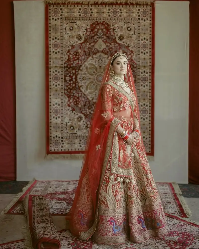 Full-length portrait of a bride in a red and gold lehenga standing on layered antique rugs
