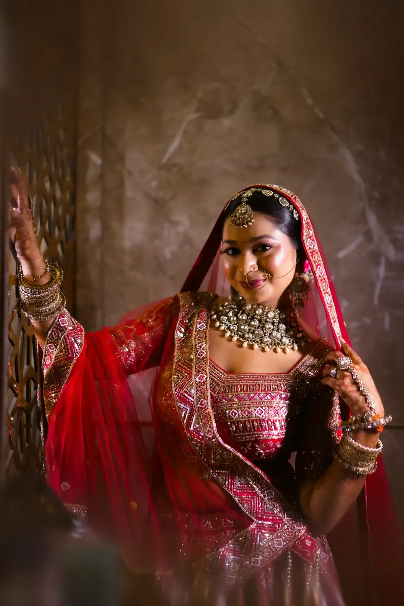 A bride smiling as she lifts her red dupatta against a marble wall