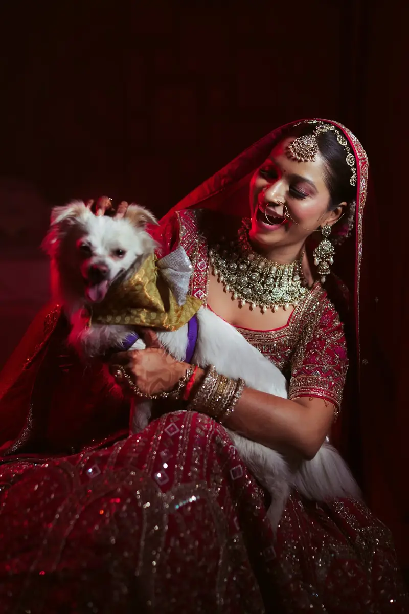 A bride in a red lehenga laughing as she holds her small white dog