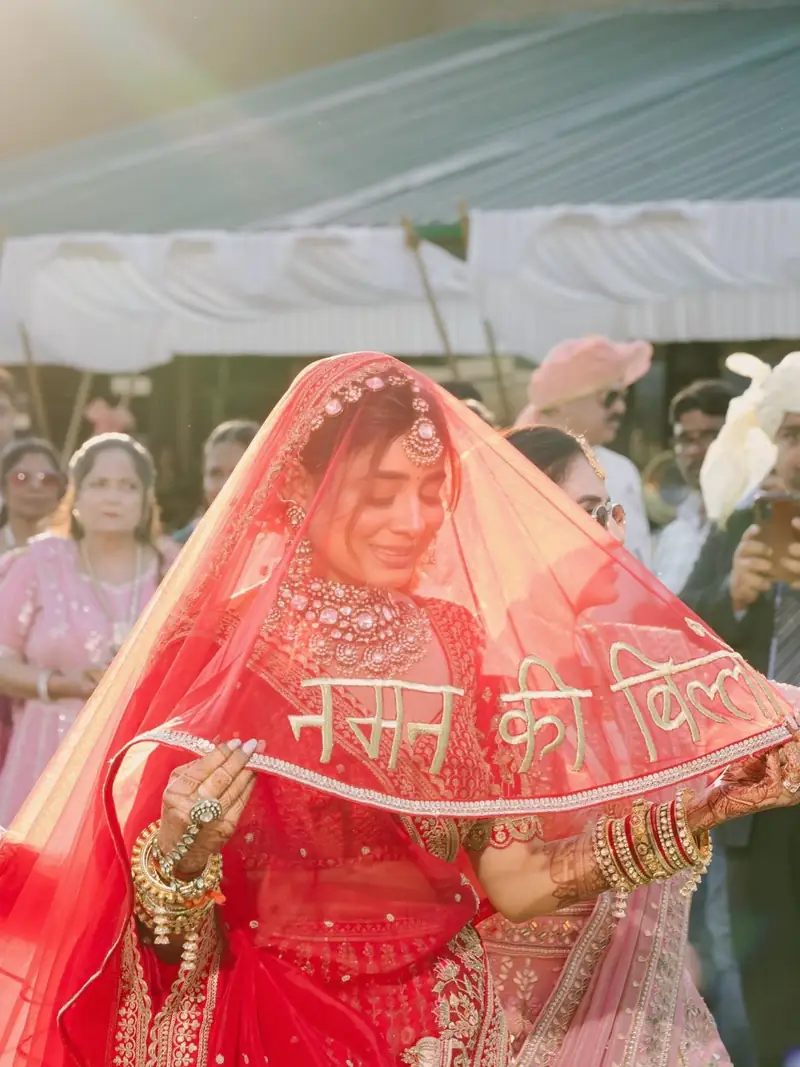 The bride smiling under a sheer red veil embroidered with Hindi words as she walks in