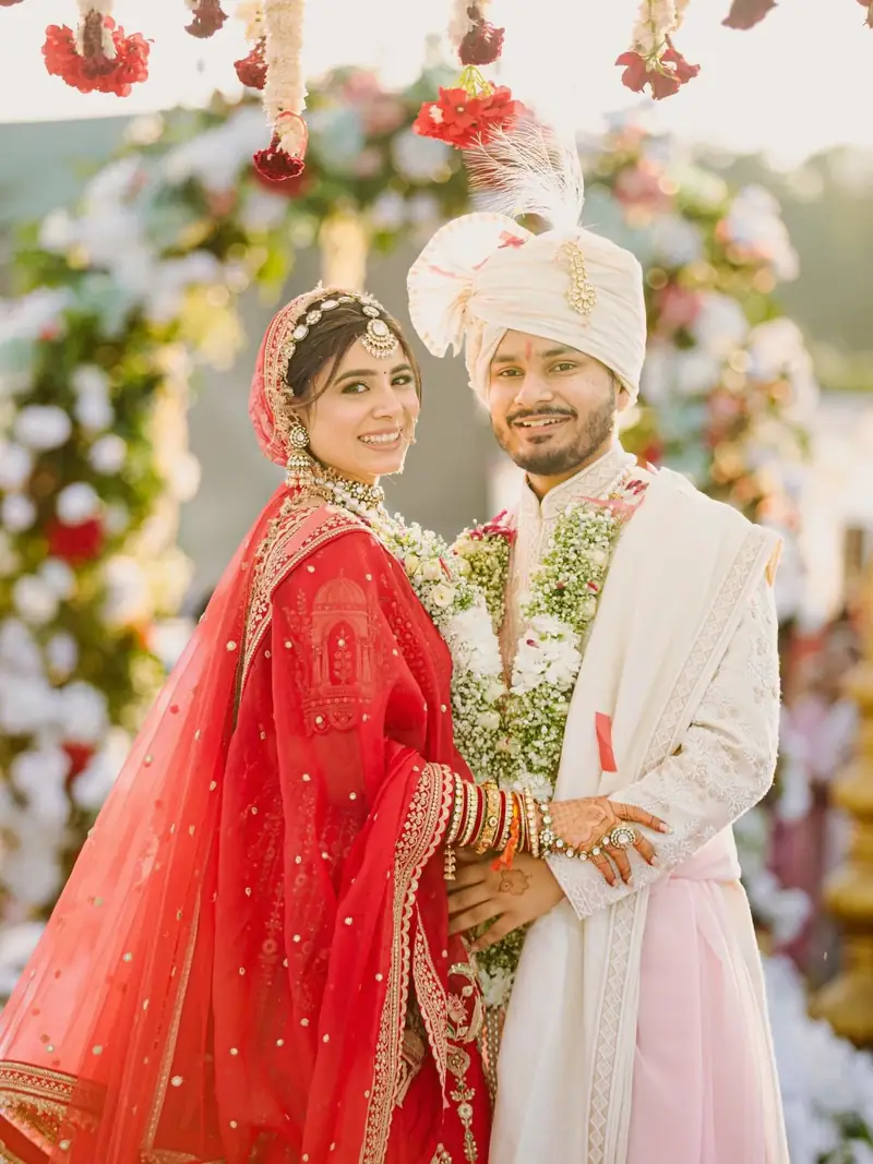 The couple holding each other and smiling, she in red, he in ivory with a pink stole