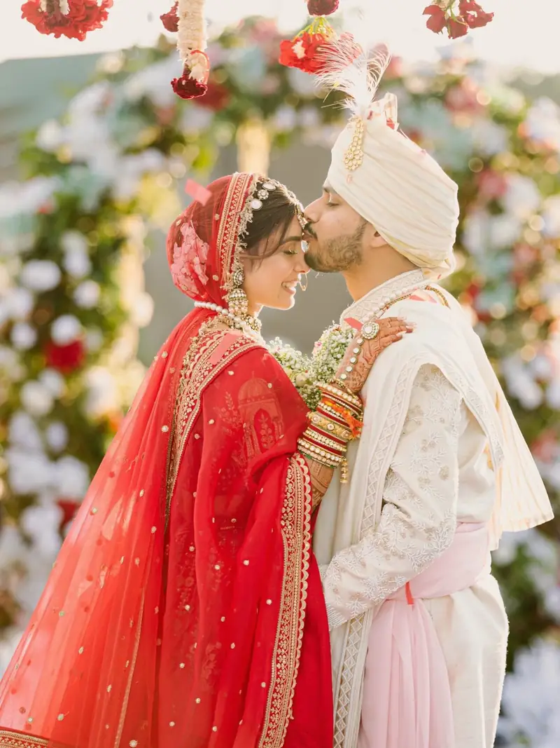 The groom kissing the bride's forehead in front of a red and white floral arch