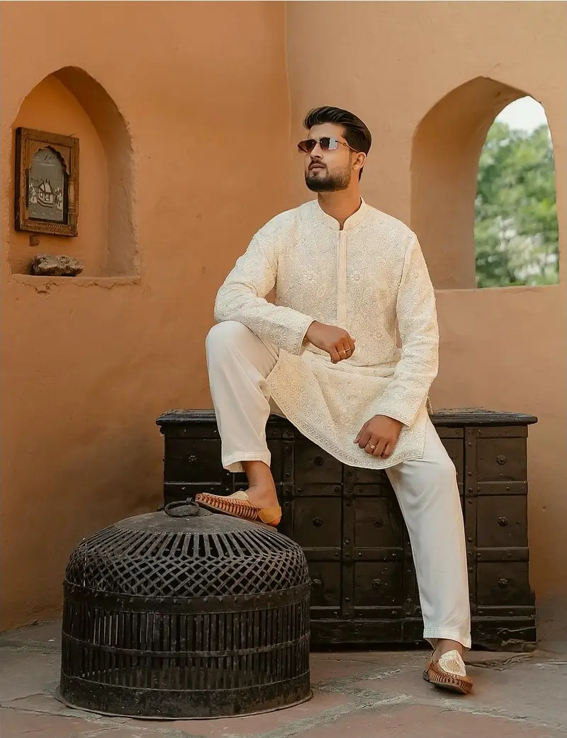 The groom in an ivory kurta and sunglasses, seated on an old wooden chest beneath arched windows
