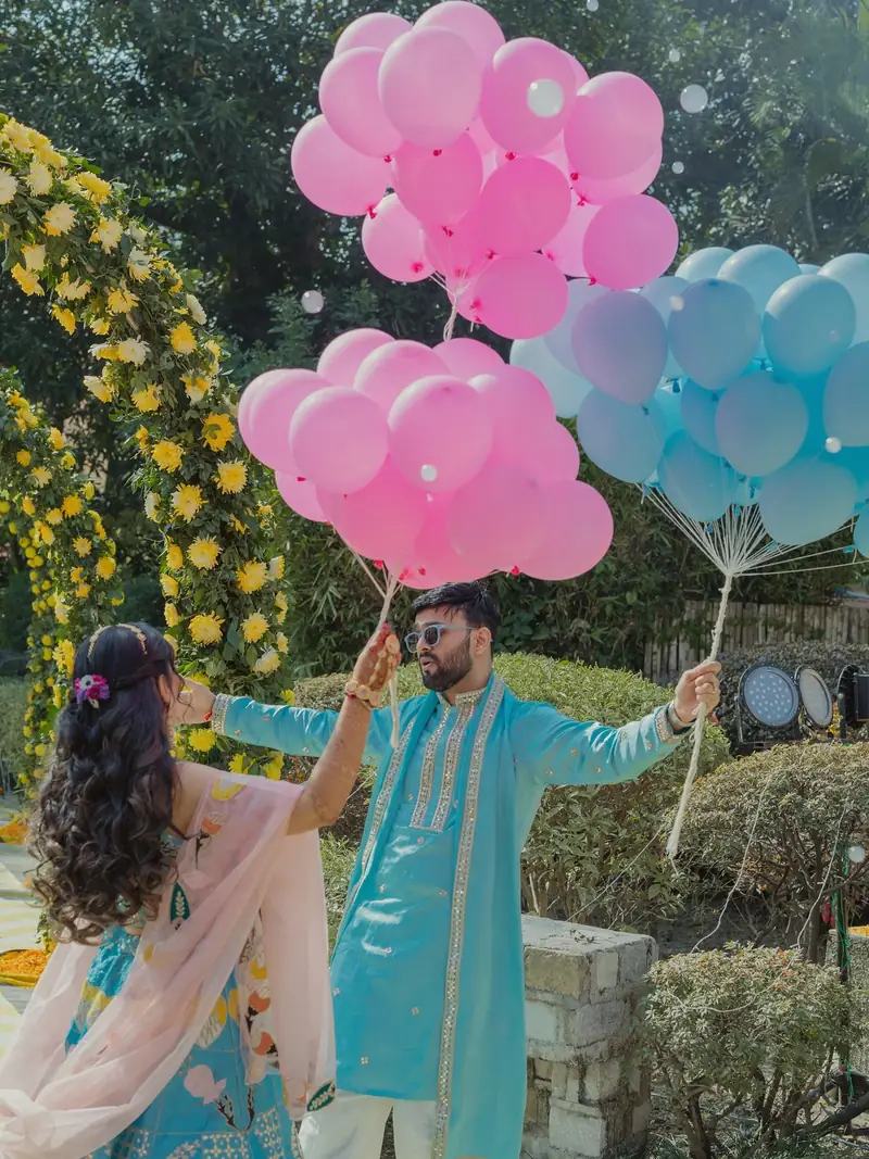 The couple holding bunches of pink and blue balloons beside a marigold arch