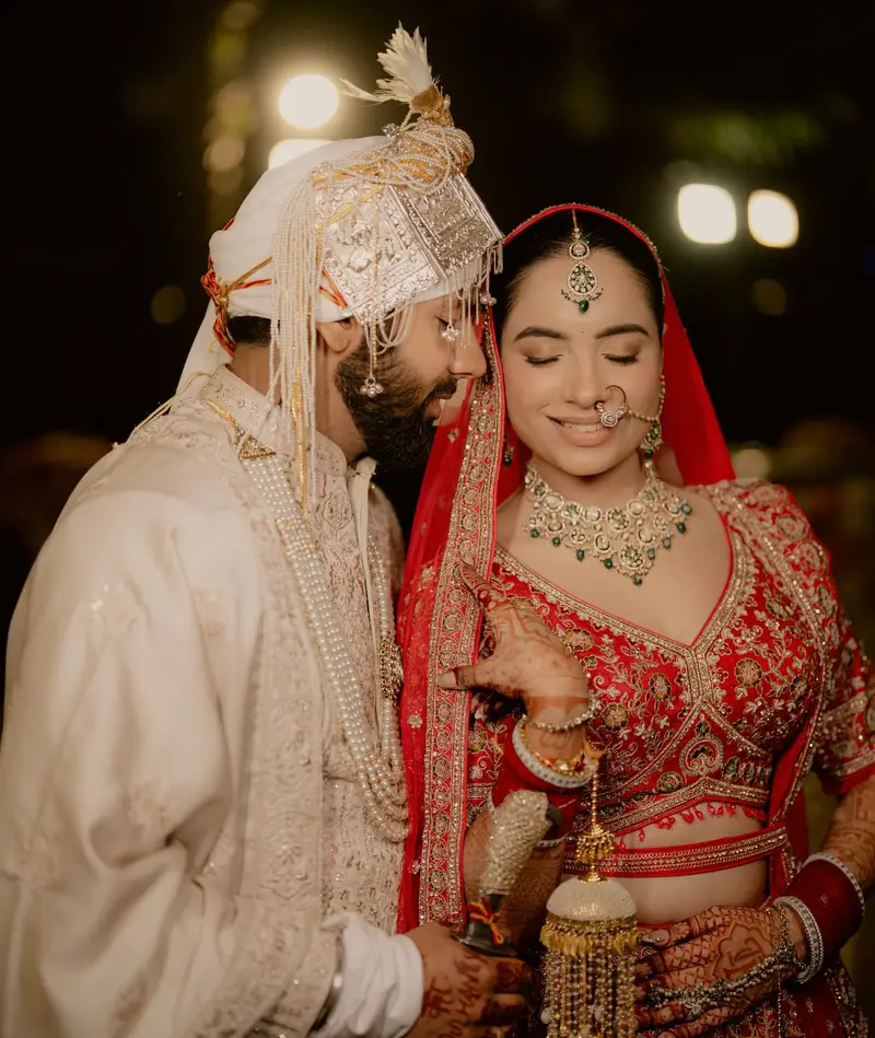 The bride smiling at the camera as the groom leans in close, warm lights behind them
