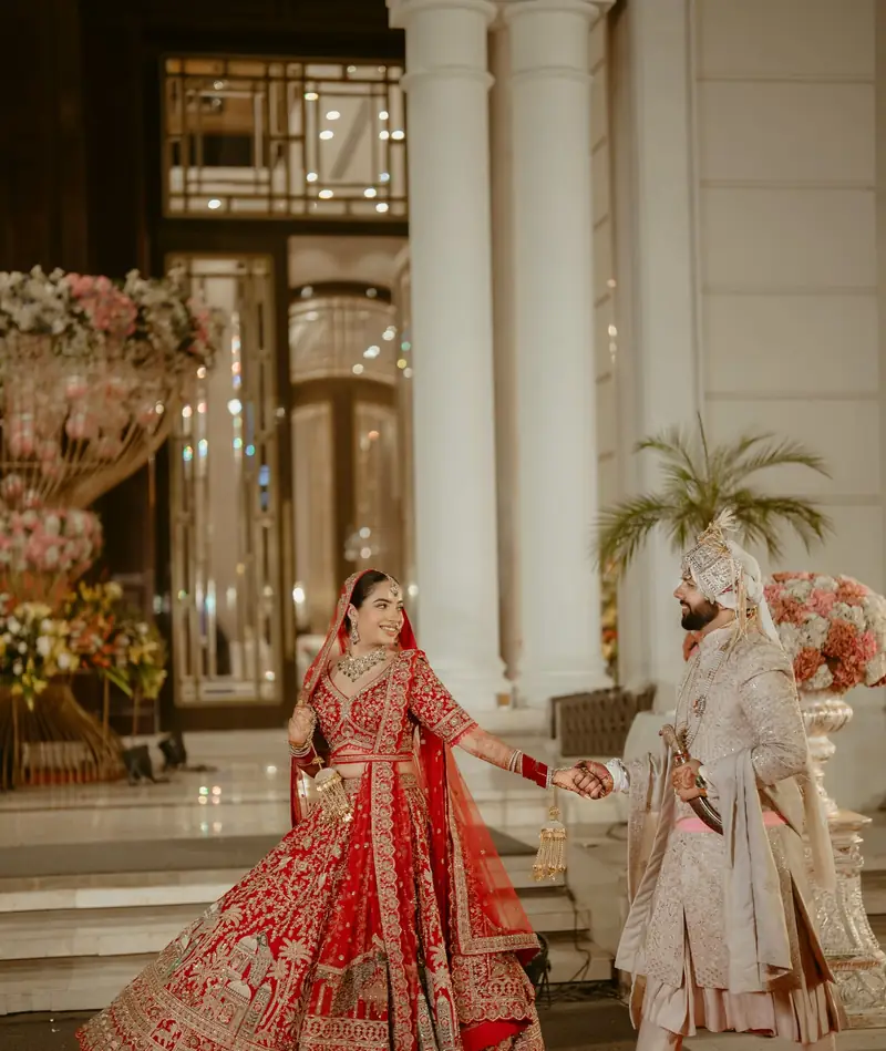 The bride twirling her red lehenga as the groom walks towards her between white columns