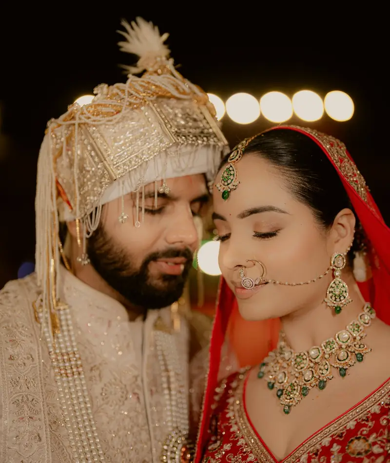 Close portrait of the couple with their eyes closed, her nath and his sehra catching the light