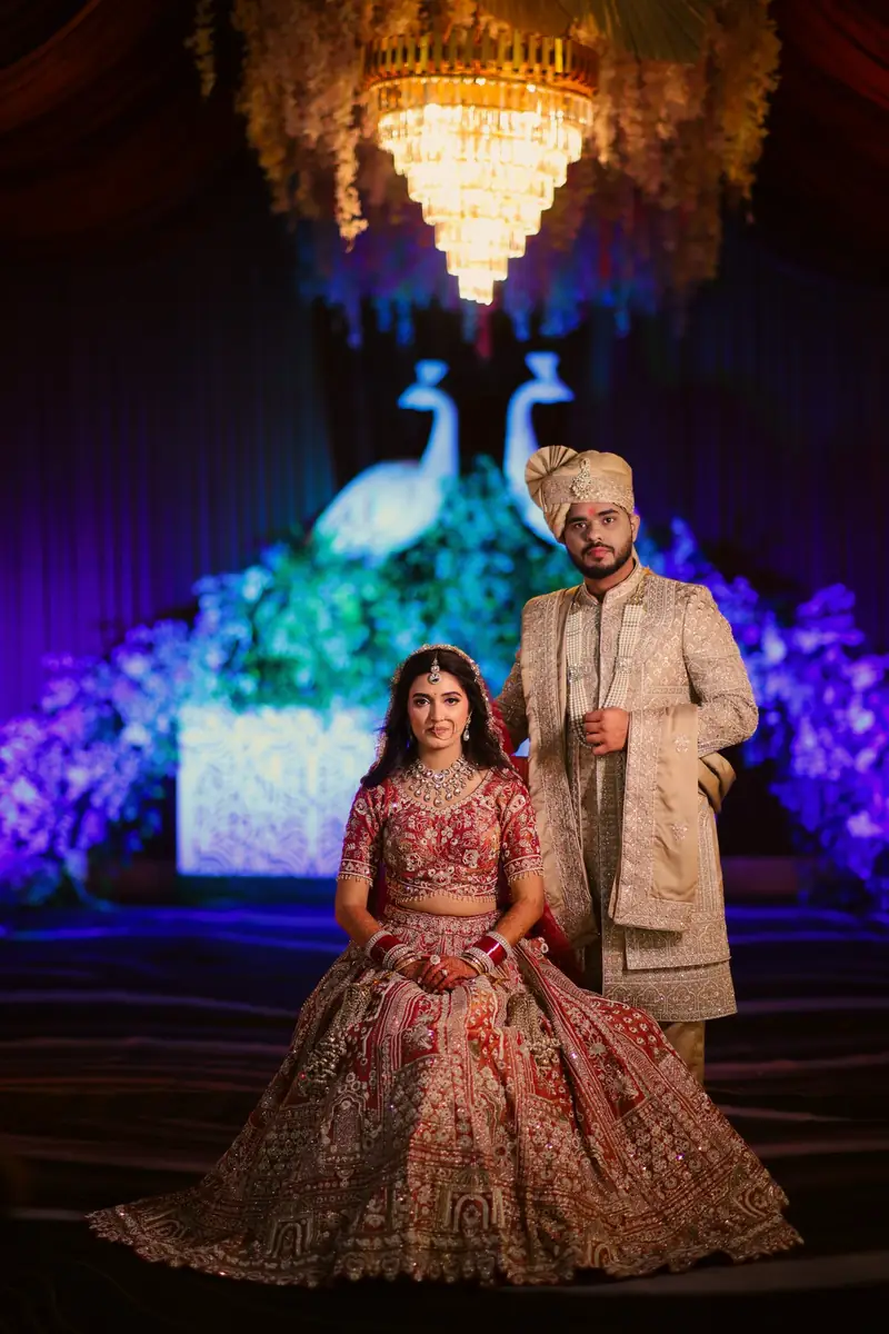 A couple on their reception stage in front of a glowing peacock backdrop and chandelier