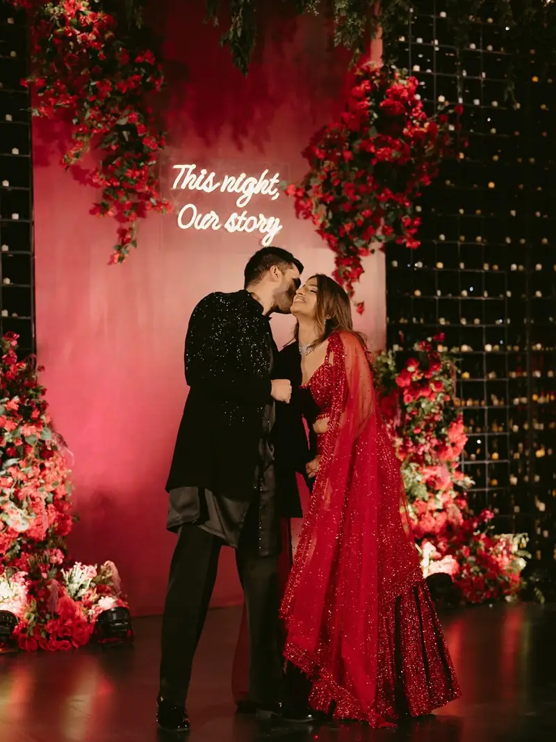 A couple sharing a kiss in front of a red floral backdrop with a neon sign reading "This night, our story"