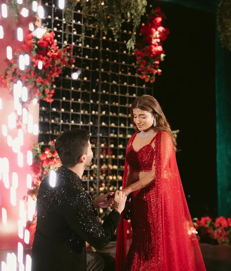 The groom on one knee holding the bride's hand beside a wall of lights