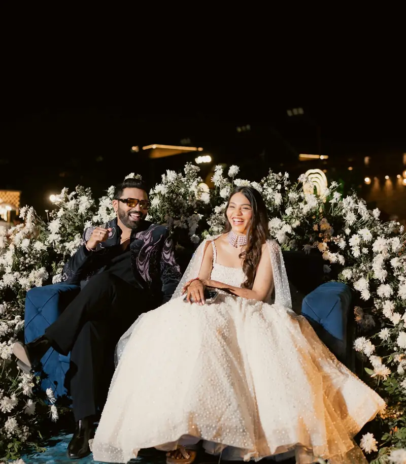 The couple laughing on a blue sofa set into a wall of white flowers