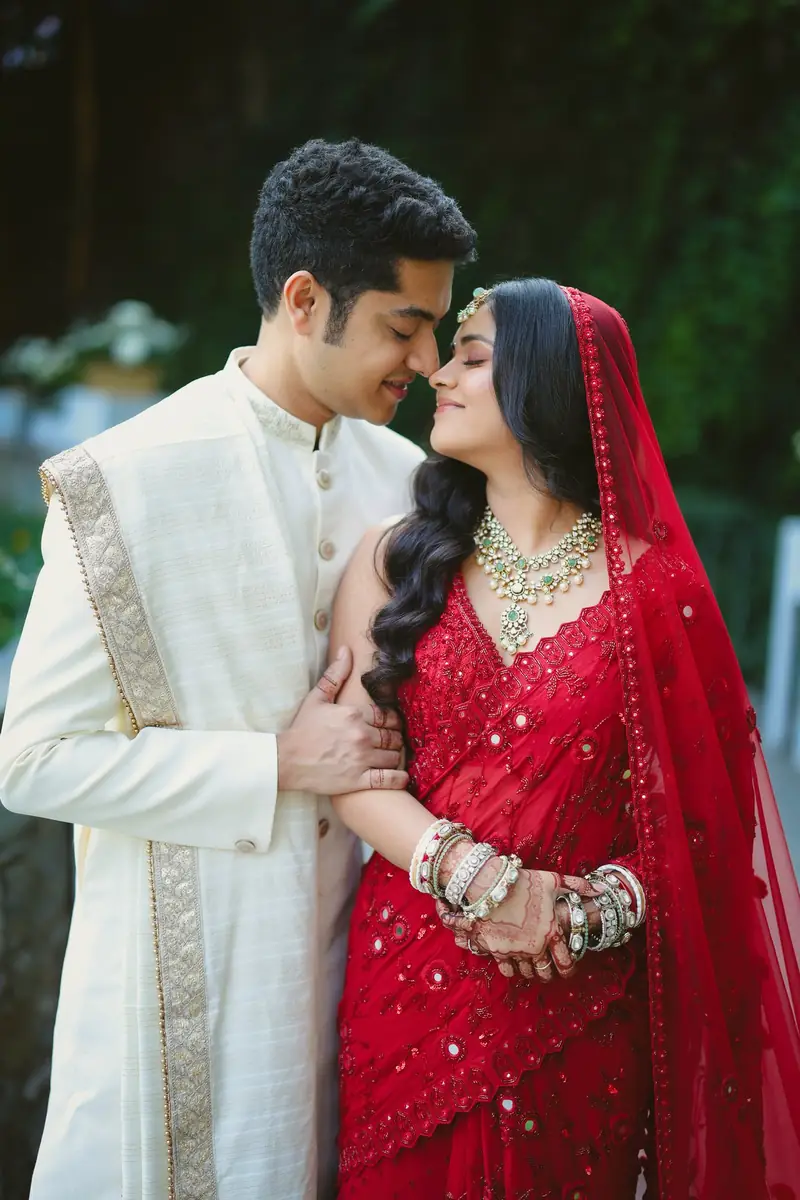 The groom in an ivory sherwani and the bride in a red saree, nose to nose in a garden