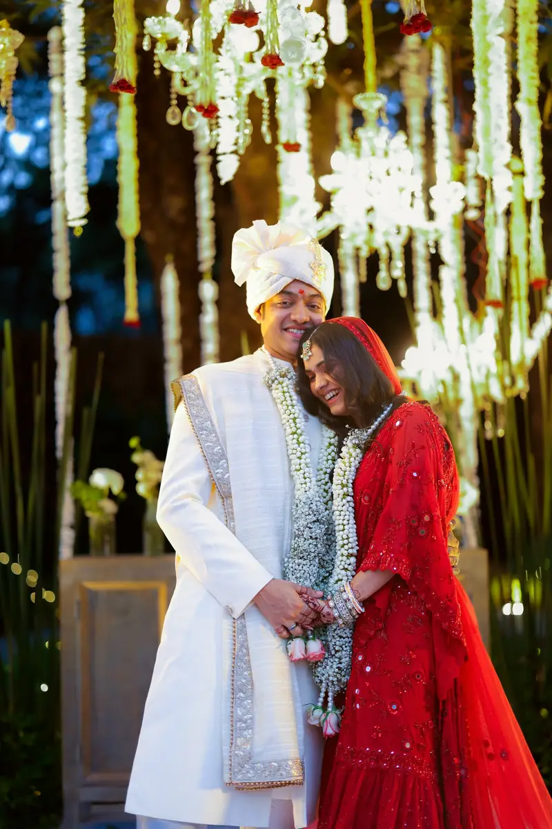 The couple in their varmala garlands, the bride leaning on the groom under hanging white flowers
