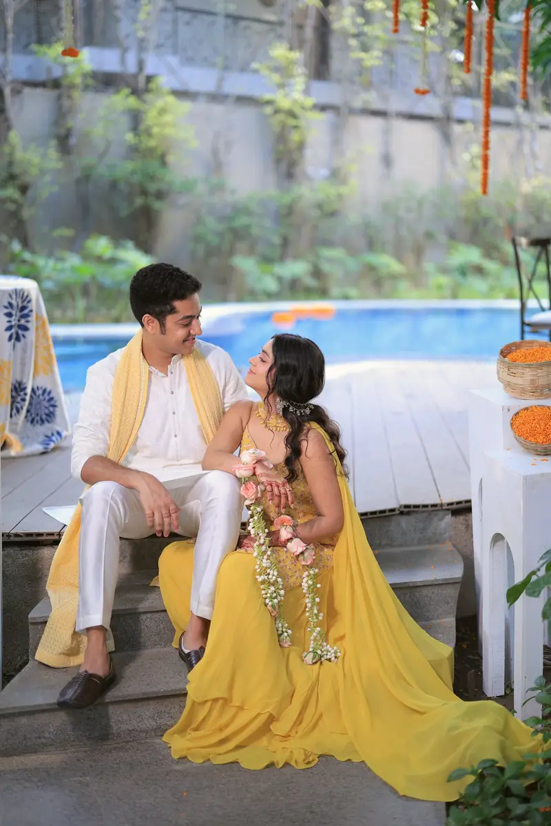 The couple sitting by the pool, she in a flowing yellow lehenga with floral jewellery