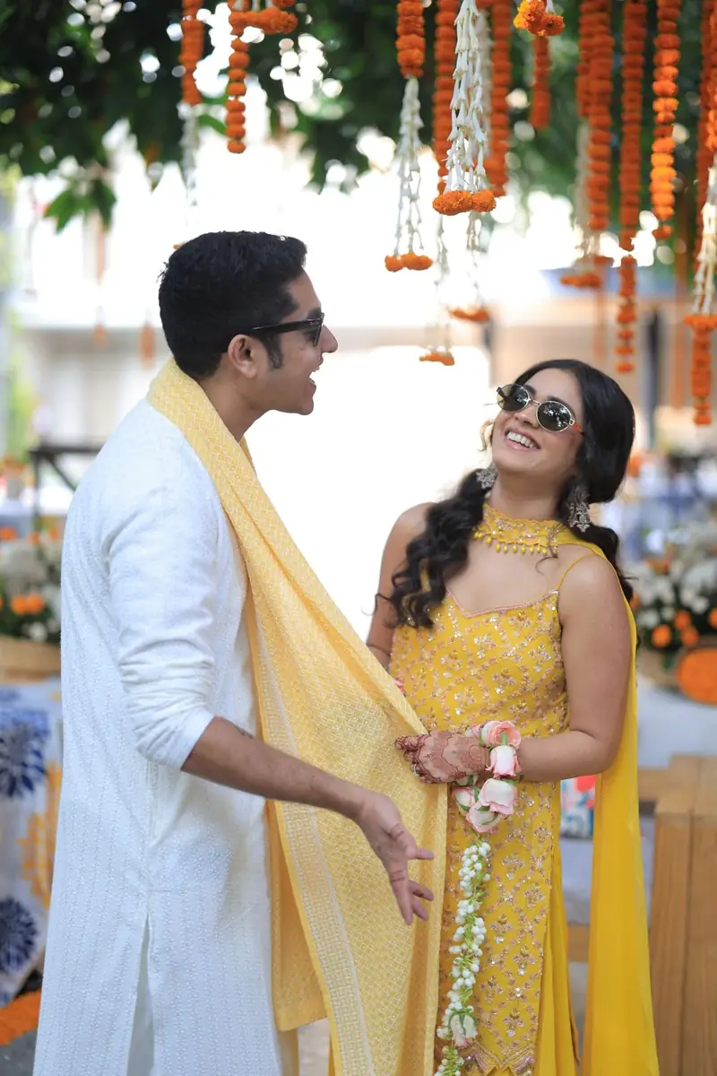 A couple laughing together at their haldi under strings of marigold